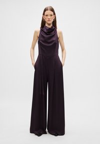 Jumpsuit bez rukávů z hlubokého fialového saténového materiálu, s drapovaným výstřihem, širokým nohavicovým střihem a bočními kapsami. Hladká textura po celém povrchu.