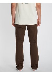 Homme portant un pantalon marron à jambes droites, un T-shirt blanc et des baskets blanches, vu de dos sur un fond uni.