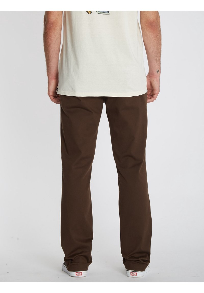 Homme portant un pantalon marron à jambes droites, un T-shirt blanc et des baskets blanches, vu de dos sur un fond uni.