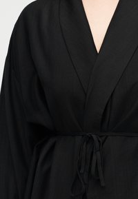 Κοντινή λήψη ενός ατόμου που φοράει μαύρη ρόμπα με φαρδιές πέτο και δεμένη ζώνη στη μέση, εμφανίζοντας το πάνω μέρος του κορμού.