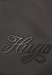 Tessuto nero testurizzato con logo "Hugo" in rilievo in un font corsivo, che crea un elemento di design sottile ma distinto.