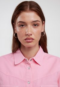 Rosa Jeanshemd mit Kragen, Druckknöpfen und Fronttaschen. Das Model trägt Ohringe und hat ein neutrales Make-up, das die natürliche Hautstruktur zur Geltung bringt.