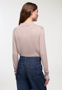 Kobieta z długimi brązowymi włosami nosi beżowy sweter polo z długim rękawem i dżinsy w kolorze niebieskim z wysokim stanem, stoi na tle jednolitego jasnego tła.