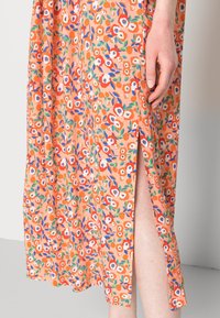 Robe florale avec un fond orange vif ornée de fleurs blanches et bleues. Fente latérale qui met en valeur le fluide du tissu léger.