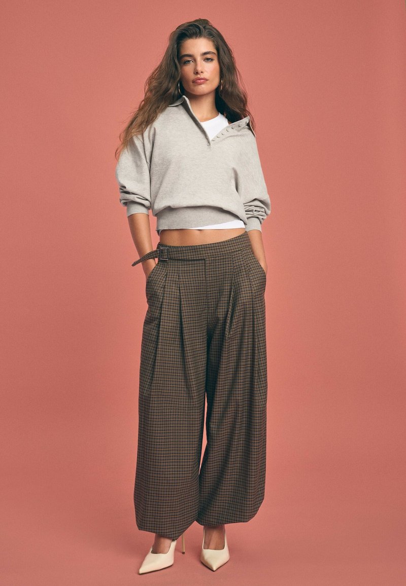 Femme posant avec un pull court beige, un débardeur blanc, un pantalon large marron à carreaux avec ceinture, et des talons hauts blancs pointus sur fond rose.