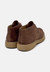 Stivaletti in suede marrone, con una texture liscia, punta rotonda e suola in gomma. Presentano tre occhielli per l'allacciatura e un dettaglio di branding sottile.