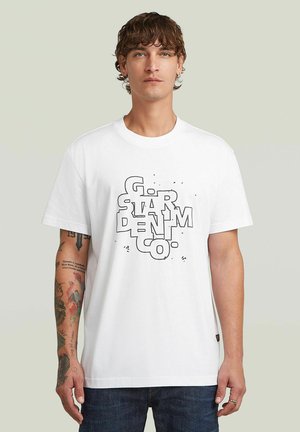 Mężczyzna z kręconymi brązowymi włosami, ubrany w białą koszulkę z grafiką z czarnym napisem "G-STAR DENIM CO." oraz ciemne dżinsy, stojący na jednolitym tle.