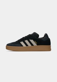 Black suede sneaker with three white strip accents, cushioned collar, and a brown rubber sole featuring a textured grip pattern.