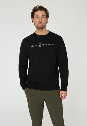 Sudadera negra con el logo "SAIL RACING" en blanco, cuello redondo, puños y dobladillo acanalados. Combinada con pantalones verde oliva.
