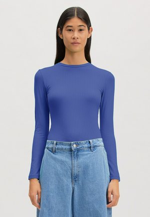 Frau mit langen dunklen Haaren, die ein eng anliegendes blaues geripptes Langarmshirt und hoch taillierte hellblaue Jeans vor schlichtem Hintergrund trägt.