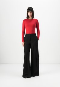 Vrouw met steil zwart haar draagt een rode lange mouw top, zwarte wijde cargo broek en zwarte schoenen, staande tegen een witte achtergrond.
