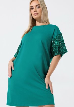 Blonde Frau in locker sitzendem, grünem Kleid mit kurzen Ärmeln, verziert mit grünen Pailletten, vor weißem Hintergrund stehend.