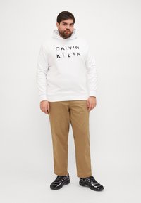 Biały kaptur z napisem "CALVIN KLEIN" w czarnym kolorze, zestawiony z beżowymi chinosami i czarnymi butami sportowymi z różowymi akcentami. Jednolity tło.