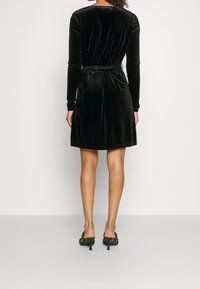 Svart sammetsklänning med långa ärmar och cinchad midja. Slät textur, knälång fåll och en enkel design. Bärs med öppen tå-hälar.