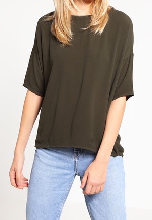 Lyshåret person iført løs mørke olivengrøn kortærmet top og lyseblå højtaljede jeans, stående mod en hvid baggrund.