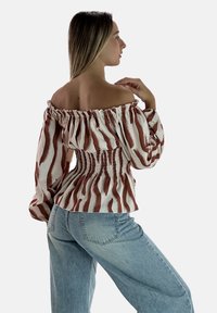 Biała bluzka z odkrytymi ramionami i marszczonym paskiem w talii, ozdobiona brązowymi falistymi wzorami. Długie rękawy z elastycznymi mankietami. W połączeniu z jasnymi dżinsami.