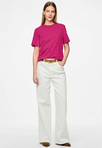 Femme debout portant une chemise à manches courtes rose vif, un pantalon blanc taille haute à jambes larges, une ceinture marron et des chaussures pointues marron.