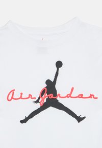 Σιλουέτα παίκτη μπάσκετ που πηδάει με τη μπάλα πάνω από το κεφάλι, κόκκινο κείμενο "Air Jordan" πάνω σε λευκό μπλουζάκι.