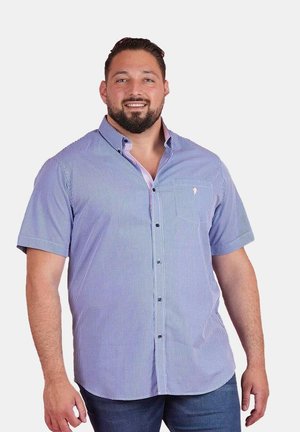 Homme souriant, portant une chemise à manches courtes bleu clair à carreaux et un pantalon bleu foncé, debout devant un fond blanc uni.