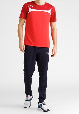 Röd idrottströja med vita detaljer, tillsammans med marinkolored mjukisbyxor. Har en logotyp på både tröjan och byxorna, samt vita sneakers.