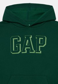 Haut à capuche vert en tissu doux arborant un grand logo "GAP" en vert clair. Il possède une capuche classique et une poche kangourou.