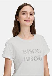 Hvid bomulds t-shirt med oprullede ærmer, der har teksten "BISOU BISOU" i grå broderet skrift på forsiden. Blød tekstur, afslappet pasform.