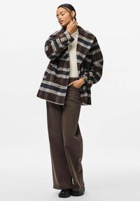 Cappotto a scacchi marrone e blu con colletto a punte, chiusura a bottoni, abbinato a pantaloni larghi marroni e calzature nere. Texture liscia.