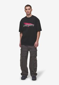 Black oversized t-shirt with white and pink logo, paired with grey cargo pants featuring multiple pockets and a relaxed fit. Black sneakers.