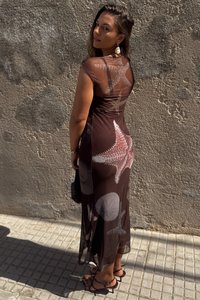 Vestido maxi em tecido translúcido castanho com padrões de estrelas-do-mar e conchas. Apresenta um corpete justo e uma saia fluida. Usado com sandálias pretas.