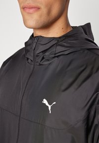 Svart lättviktsjacka med huva, med en dragkedja i full längd och en vit Puma-logotyp på bröstet. Slät yta och sportig design.
