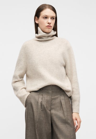 Γυναίκα που φοράει μπεζ πουλόβερ με ζιβάγκο, μπαίνει μέσα σε ψηλόμεσο, φαρδύ παντελόνι με γκρι καρό, σε απλό φόντο.