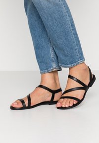 Des pieds portant des sandales plates noires à lanières, associés à un jean en denim bleu court, se tenant sur un sol de couleur claire.