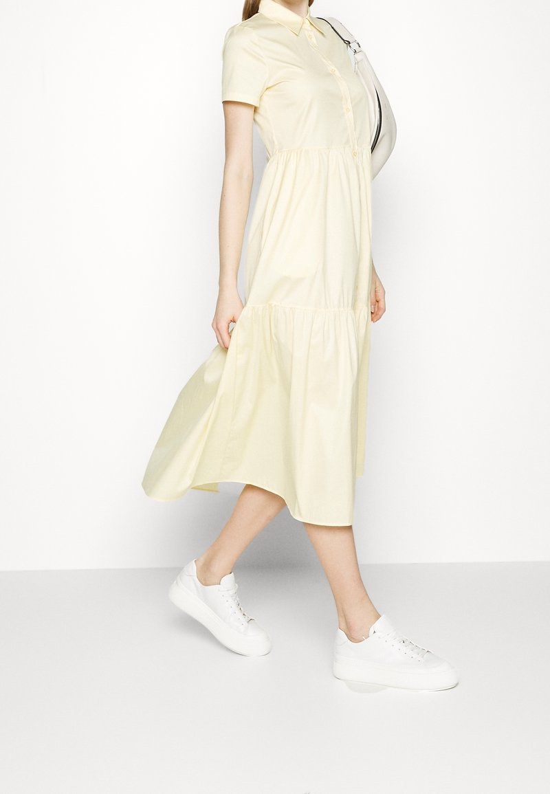 Personne marchant dans une robe midi jaune clair à manches courtes et des baskets blanches, portant un sac à épaule blanc.