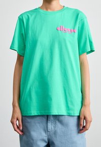 Henkilö, joka on pukeutunut löysään turkoosiin T-paitaan, jossa on vaaleanpunainen "ellesse"-logo rintamalla, ja vaaleansinisiin housuihin, yksinkertaista taustaa vasten.