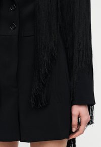 Κοντινό πλάνο ενός ατόμου που φοράει μαύρο, κουμπωμένο παλτό με μακριά μαύρη φούντα και το δεξί του χέρι χαλαρά δίπλα στο σώμα.