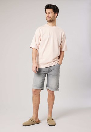 Man staat met één hand in zijn zak, draagt een lichtroze oversized T-shirt, grijze opgerolde korte broek en beige instapschoenen.