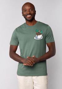 Grünes Baumwoll-T-Shirt mit gesticktem Design eines Bechers mit Bergen und Palmen. Weicher Stoff, Rundhalsausschnitt, kurze Ärmel.