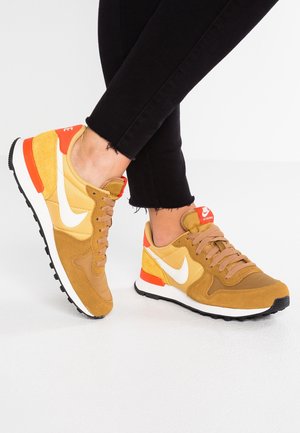 Fødder iført Nike sneakers i sennepsgul og orange med hvid swoosh, kombineret med sorte korte bukser mod en hvid baggrund.