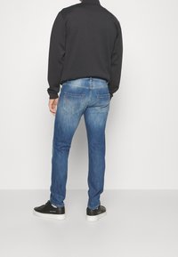 Tmavý svetr, modré džínové kalhoty s opotřebovaným vzhledem, obsahující zadní kapsy a detaily se zipem, kombinované s černými teniskami.