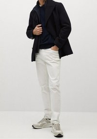 Cappotto in misto lana navy su un maglione navy, pantaloni bianchi slim-fit e sneakers grigio chiaro con dettagli scuri; outfit casual a strati.