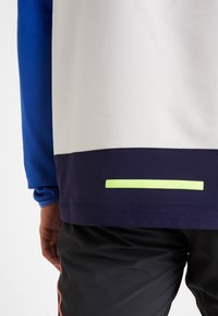 Koszulka sportowa z krótkim rękawem, z niebieskimi siateczkowymi rękawami, białym tułowiem oraz granatowym obszyciem z odblaskowym żółtym paskiem na dole.