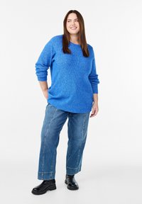Kobieta uśmiechająca się, ubrana w jasnoniebieski sweter, szerokie dżinsy i czarne masywne botki, stojąca na białym tle.