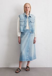 Lichtblauwe denimvest met knopen en zakken over een crèmekleurige blouse, gecombineerd met een bijpassende denimrok. Zwarte sandalen maken de outfit compleet.