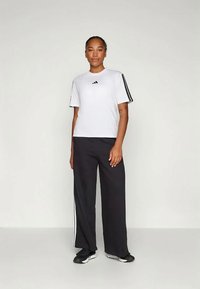 T-shirt branca de manga curta com logótipo preto e calças de treino pretas com riscas laterais brancas. O material parece leve e respirável.