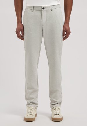 Man draagt lichtgrijze, gestructureerde broek met zwarte knoop en off-white sneakers met gumzolen, staand tegen een effen achtergrond.