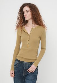 Γυναίκα με μακριά σγουρά κόκκινα μαλλιά, που φοράει μία μουσταρδί ριμπ μπλούζα henley με μακριά μανίκια και φαρδιά μπλε τζιν, στέκεται μπροστά σε απλό λευκό φόντο.