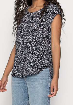 Vrouw draagt een zwart topje met korte mouwen met een kleine witte bloemenprint en lichtblauwe spijkerbroek, zichtbaar van nek tot heupen.