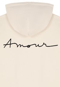 Kremeni dukserica s prednjim džepom, koja na gornjem dijelu leđa sadrži crnu vezenu reč "Amour" u kurzivnom fontu.
