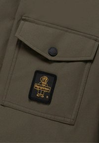 Tessuto verde oliva con tasca a patta, caratterizzato da una toppa rettangolare nera con una grafica di un cane giallo e il testo "RefrigiWear".