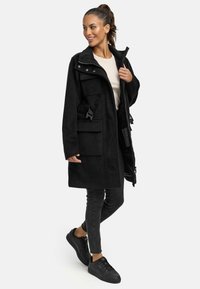 Cappotto in pile nero con colletto alto, chiusura con zip, tasche frontali e cinturini regolabili. Abbinato a jeans skinny neri e sneakers.